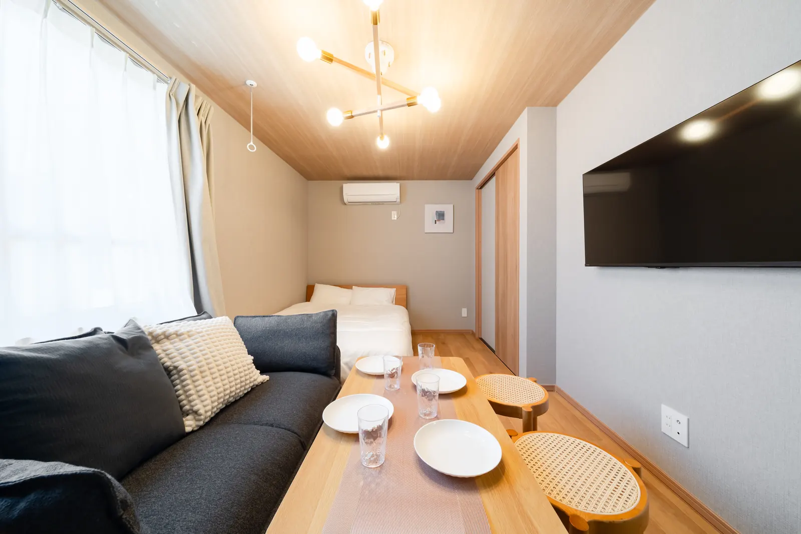 Living side of a studio unit at Intervia Aoto: navy sofa, wooden dining table with stools, double bed and pendant light